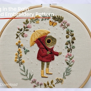 May include: Hand embroidery pattern featuring a frog in a red coat and yellow boots holding a yellow umbrella. The frog is surrounded by a floral wreath with butterflies and the text "Frog in the Rain Hand Embroidery Pattern".