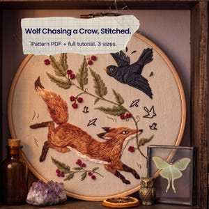 Op de afbeelding: Een borduurring met een geborduurd ontwerp van een vos die naar een zwarte kraai rent tussen groen gebladerte en rode bessen. De tekst op de ring luidt: Wolf Chasing a Crow, Stitched. Pattern PDF + full tutorial. 3 sizes.