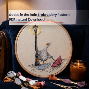 May include: An embroidery hoop featuring a stitched design of a white goose holding a pink umbrella while standing by a black lamppost in the rain. The text Goose in the Rain Embroidery Pattern PDF Instant Download is visible above the hoop.