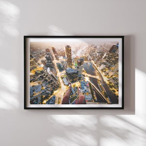 Puede incluir: Fotografía aérea enmarcada de un paisaje urbano al anochecer. La imagen captura una vista de ángulo alto de edificios, calles y un río iluminados. La perspectiva incluye las piernas y los pies de una persona, añadiendo una sensación de escala y aventura.