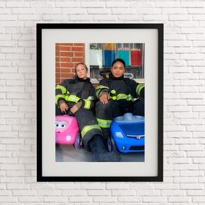 May include: A framed photograph featuring two firefighters in dark uniforms with reflective yellow stripes. They are seated, each with a toy car in front of them, one pink and one blue. The background is a colorful building.
