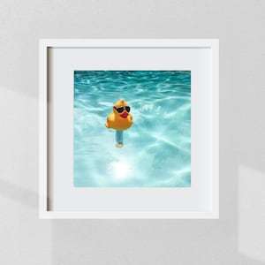 以下が含まれることがあります： サングラスとデニムショーツを身に着けた黄色のラバーダックが、透明でターコイズブルーの水の中で浮かんでいるフレームプリント。アヒルは焦点であり、さざ波の立つ水と太陽光を背景にしています。