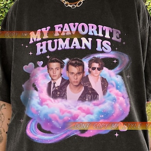 Könnte beinhalten: Schwarzes T-Shirt mit dem Text "MY FAVORITE HUMAN IS" und einer Grafik von drei Männern in einer bunten Wolke. Das T-Shirt hat einen Vintage-Look.