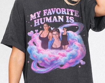 Puede incluir: Camiseta gris oscuro con el texto "MY FAVORITE HUMAN IS" en un degradado de rosa y morado. La camiseta presenta un gráfico de tres personas dentro de una nube de colores rosa, azul y morado.