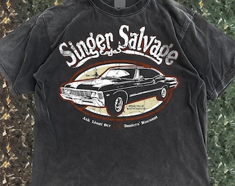 Puede incluir: Camiseta gris oscuro con un diseño de coche clásico dentro de un marco ovalado. El texto "Singer Salvage" está sobre el coche, con "Sioux Falls, South Dakota" y "Ask About Our Hunters' Discount" debajo. Estilo vintage.