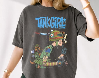 Puede incluir: Camiseta gris oscuro con el texto "TANK GIRL" en azul. El gráfico presenta una ilustración de dibujos animados de una persona con uniforme militar, con un casco y un dedo en los labios. El diseño incluye acentos verdes, rojos y amarillos.