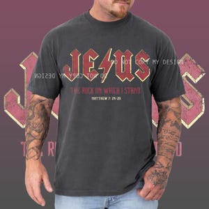 Op de afbeelding: Donkergrijs t-shirt met het woord "JESUS" in rode en gouden letters. De tekst "THE ROCK ON WHICH I STAND" en "MATTHEW 7:24-25" staan eronder. Het shirt heeft korte mouwen en een vintage stijl.