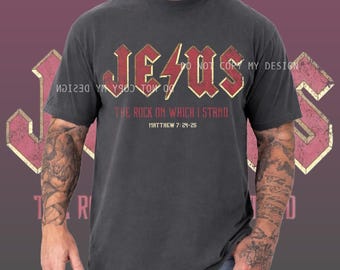 Puede incluir: Camiseta gris oscuro con la palabra "JESUS" en rojo y dorado. Debajo, el texto "THE ROCK ON WHICH I STAND" y "MATTHEW 7:24-25". La camiseta tiene mangas cortas y un estilo vintage.