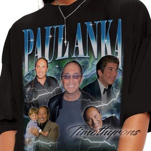 Puede incluir: Camiseta negra con un gráfico de Paul Anka, con múltiples imágenes de él, el nombre "PAUL ANKA" en azul y blanco y el texto "Timothyrons". El diseño tiene una estética vintage con acentos de rayos.