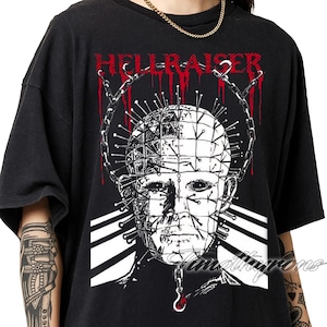 May include: Black t-shirt featuring a graphic of a character from the horror film "Hellraiser." The design includes a detailed illustration of the character's face, a crown of nails, and the word "HELLRAISER" in red. The shirt is a classic crew neck style.