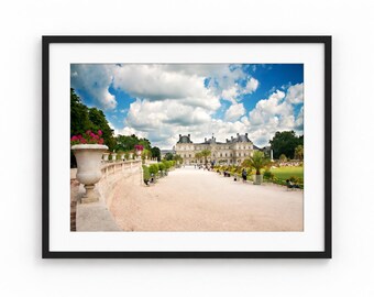 May include: A framed landscape photograph of the Luxembourg Palace in Paris, featuring a wide gravel path, stone balustrades with large planters filled with pink flowers, green trees, and a bright blue sky with white clouds above the building.