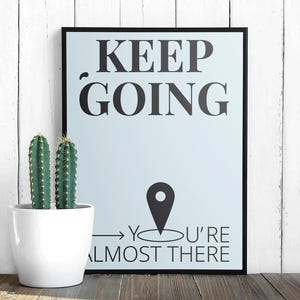 Puede incluir: Un póster enmarcado con las palabras "KEEP GOING" en negro, con un icono de ubicación y el texto "YOU'RE ALMOST THERE". Una maceta blanca con dos cactus verdes está frente al póster. El fondo es una pared de madera blanca.
