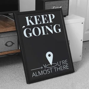 Puede incluir: Cartel enmarcado en negro con texto blanco que dice "KEEP GOING" encima de un gráfico de marcador de ubicación y las palabras "YOU'RE ALMOST THERE". Un pequeño altavoz negro se encuentra en un armario blanco en el fondo.
