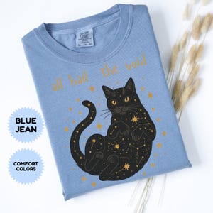 May include: Blue jean Comfort Colors t-shirt with a black cat illustration. The cat is lying on its back with a constellation design and the words "all hail the void" in gold lettering. The shirt is folded.