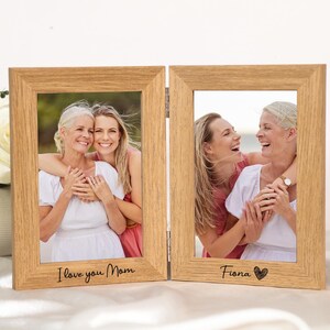 May include: A wooden, two-panel photo frame with pictures of two women. The left panel reads "I love you Mom", and the right panel reads "Fiona" with a heart symbol. The frame is made of light-colored wood.