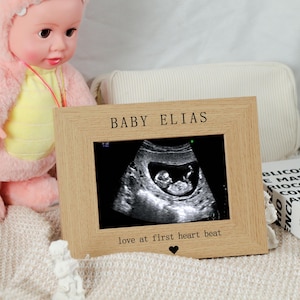 May include: A wooden picture frame with the text "BABY ELIAS" at the top and "love at first heart beat" at the bottom. The frame holds a grayscale ultrasound image of a fetus. A small black heart is below the text.