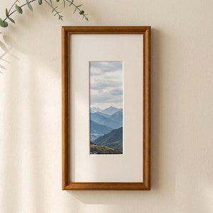 Könnte beinhalten: Ein rechteckiger Bilderrahmen aus Holz mit mittelbraunem Finish und einem breiten, cremeweißen Passepartout. Der Rahmen enthält ein vertikales Foto, das eine Landschaft aus geschichteten blauen Gebirgszügen unter einem bewölkten Himmel zeigt.