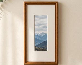 May include: A rectangular wooden picture frame with a medium brown finish and a wide off-white mat board. The frame contains a vertical photograph depicting a landscape of layered blue mountain ranges under a cloudy sky, centered within the mat.