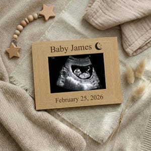 May include: A light-coloured wooden picture frame with the text "Baby James" and a crescent moon. The frame holds a black and white ultrasound image. The date "February 25, 2026" is below the image. A wooden star mobile is in the background.