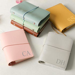 以下が含まれることがあります： ピンク、グレー、イエロー、グリーン、ブラウンなど、さまざまなパステルカラーのパーソナライズされた革製ジャーナルのコレクション。各ジャーナルにはイニシャルとゴムバンドが付いています。ジャーナルは幅約10cm、高さ約15cmです。