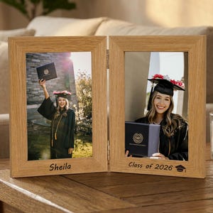 May include: A light-coloured wooden hinged photo frame with two graduation photos. The left frame has the name "Sheila" and the right frame says "Class of 2026". The photos show a graduate in a cap and gown.