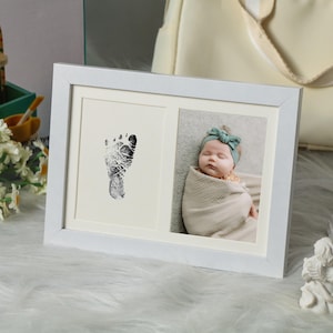 May include: A white double picture frame. One side features a black footprint, the other a baby wrapped in a blanket, wearing a green headband. The frame is on a fluffy white surface.