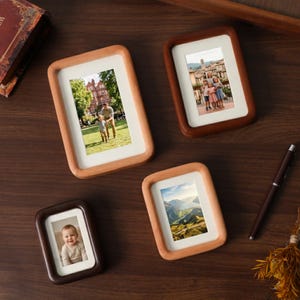 Puede incluir: Cuatro marcos de fotos rectangulares de madera con esquinas redondeadas, cada uno con una fotografía diferente. Los marcos son de tonos marrón claro y oscuro, con paspartús blancos alrededor de las fotos. También se ven un bolígrafo y un libro.