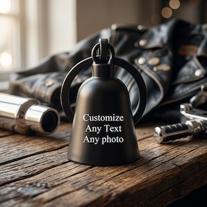 May include: A matte black bell-shaped motorcycle guardian bell with a circular attachment ring at the top. The front surface features white printed text that reads: Customize Any Text Any photo. The object has a smooth, solid finish.