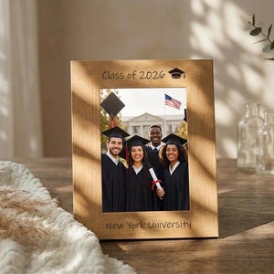 以下が含まれることがあります： 「Class of 2026」の文字と卒業帽子のアイコンが入った木製のフォトフレーム。フレームには、ガウンと帽子をかぶった4人の卒業生の写真が収められており、下部には「New York University」の文字が入っています。