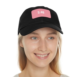 May include: Black baseball cap with a pink rectangular patch. The patch displays the time "5:46" in white. The cap is worn by a person with blonde hair and a gray t-shirt.