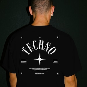 May include: Black t-shirt with the word "TECHNO" in a stylized white font. Below the word is a white starburst graphic. The shirt also features the words "BERGHAIN" and "BERLIN" and the text "ONE WHO IS THE FOCUS OF ADMIRATION THE CENTER OF ATTENTION".