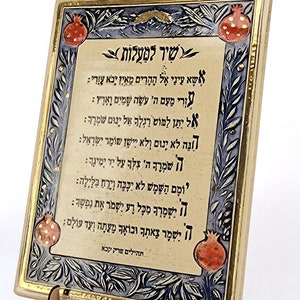 以下が含まれることがあります： 金色の枠と青と白の花柄が施された装飾的なセラミック製のプレート。プレートには、黒と赤で書かれたヘブライ語の祈り「シール・ラマアロット」が書かれています。プレートには、金色のアクセントが施された赤いザクロが2つ飾られています。