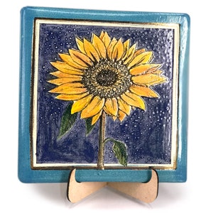 May include: A ceramic tile with a blue border and a yellow sunflower with a dark center on a blue background with white dots. The tile is on a wooden stand.