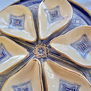 Puede incluir: Un plato de servir de cerámica blanca con cinco compartimentos individuales. El plato está decorado con un patrón geométrico azul y dorado. El centro del plato presenta una estrella de David dorada.