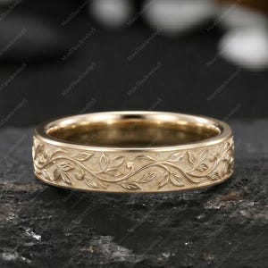May include: A gold-toned metal ring featuring a continuous engraved pattern of delicate, intertwining leaves and vines wrapping around the entire outer band. The surface of the ring has a matte finish, highlighting the intricate botanical details.