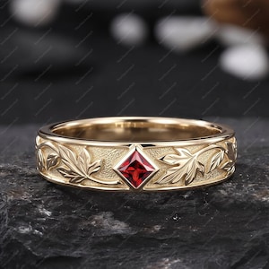 May include: A gold ring with a square, red gemstone at the center. The band features a detailed leaf pattern. The ring is displayed on a dark, textured surface, highlighting its intricate design and craftsmanship. A piece of jewelry.