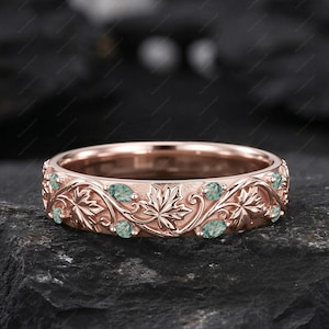 May include: Rose gold ring with a detailed leaf and vine pattern. The ring features small, light green gemstones set within the design. The ring is displayed on a dark, textured surface.