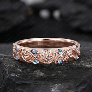 May include: Rose gold ring with a detailed leaf and vine pattern. The ring is adorned with small, oval-shaped blue gemstones. The design features intricate carvings of leaves and swirling vines, creating a nature-inspired aesthetic. The ring sits on a dark, textured surface.