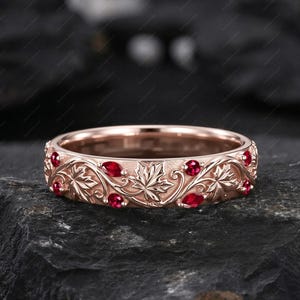 May include: Rose gold ring with a detailed leaf and vine design, accented with oval-cut red gemstones. The ring's band features an intricate pattern of leaves and curves, creating a nature-inspired aesthetic. The gemstones are evenly spaced around the band.