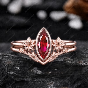Könnte beinhalten: Ein roségoldenes Ring-Set mit einem marquiseförmigen, tiefroten Edelstein. Der Ring ist mit detaillierten Ahornblatt-Designs versehen. Das Band hat ein zweilagiges Design. Der Ring ist vor einem dunklen, strukturierten Hintergrund platziert.