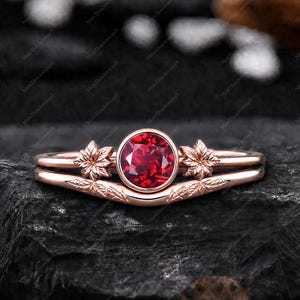 May include: A rose gold ring set featuring a round, vibrant red gemstone. The ring has a floral design on each side of the gemstone and a leaf pattern on the band. The ring is set against a dark, textured background.
