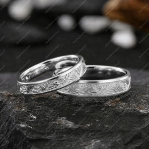May include: Two silver-toned metal rings featuring an intricate, continuous engraved leaf pattern around the outer band. The rings have a polished finish and are positioned one behind the other, showcasing the detailed botanical scrollwork design.