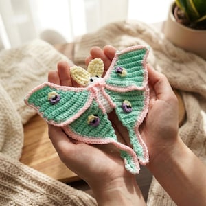 May include: A handmade crochet moth in shades of mint green, pink, and cream. The moth features detailed wing patterns and small purple accents. The wings are outlined in pink, and the body is white. A unique, handcrafted item.