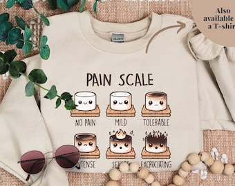 May include: Beige sweatshirt with the text "PAIN SCALE" and illustrations of marshmallows on graham crackers, each representing a level of pain from "NO PAIN" to "EXCRUCIATING". Also available as a T-shirt.