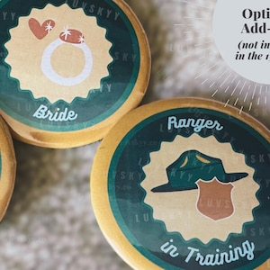 May include: Round wooden buttons with a dark green scalloped border and a tan center. One button features a white ring icon with a heart and the text Bride. Another button displays a green hat and brown badge with the text Ranger in Training.