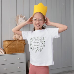 May include: A young person wearing a white t-shirt with a black and white illustration of camping gear and the text "Camp Sarah". They are wearing a yellow crown and smiling.