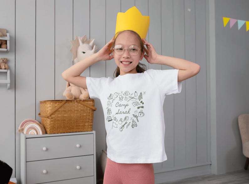 May include: A young person wearing a white t-shirt with a black and white illustration of camping gear and the text "Camp Sarah". They are wearing a yellow crown and smiling.