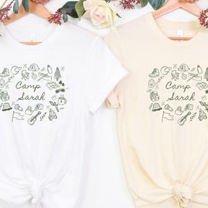 May include: Two white t-shirts with a black and green design of camping icons and the text "Camp Sarah".