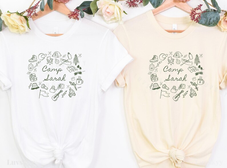 May include: Two white t-shirts with a black and green design of camping icons and the text "Camp Sarah".