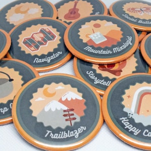May include: A collection of 10 round, colorful, illustrated buttons with a variety of nature and camping themes. The buttons feature text such as "Camp Chef", "Trailblazer", "Happy Camper", "Storyteller", "Mountain Mixologist", "Social Butterfly", "Nature's Gift", "Navigator", and "Camp Chef".